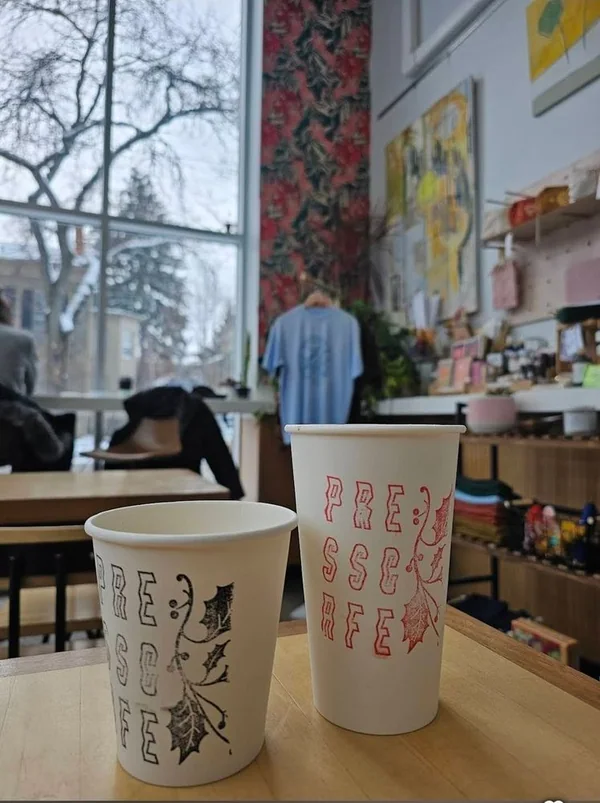 Two Press Cafe cups with interior art and local goods behind them